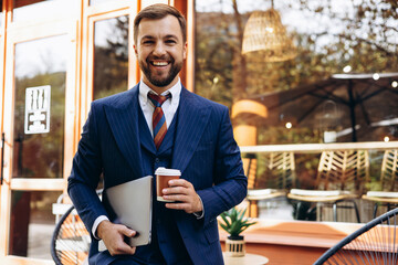 Handsome business man smiling, standing by the coffee shop holding coffee cup and his laptop