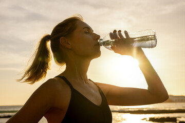 sporty woman drinking water from bottle after outdoor fitness workout at sunset