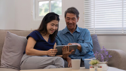 Cheerful Asian couple using network video call tablet sitting together on sofa at home Asian elderly husband sitting with elderly wife shopping online or video calling on tablet, relaxing.