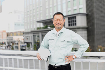 屋外のビルの前ででヘルメットを持つ作業服を着たアジア人の中年男性