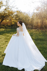 Brunette bride in a white dress with a long train, holding a bouquet and walking in the garden. Wedding photo session in nature. Beautiful hair and makeup. Celebration