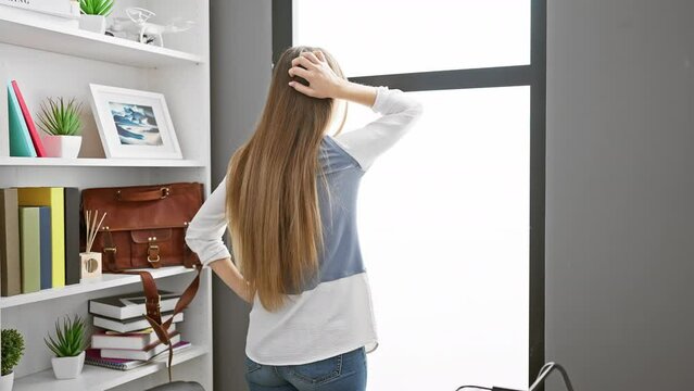 Thoughtful young blonde business woman at office, back-view of pensive worker with hand on head, expressing doubt in an indoor workplace.