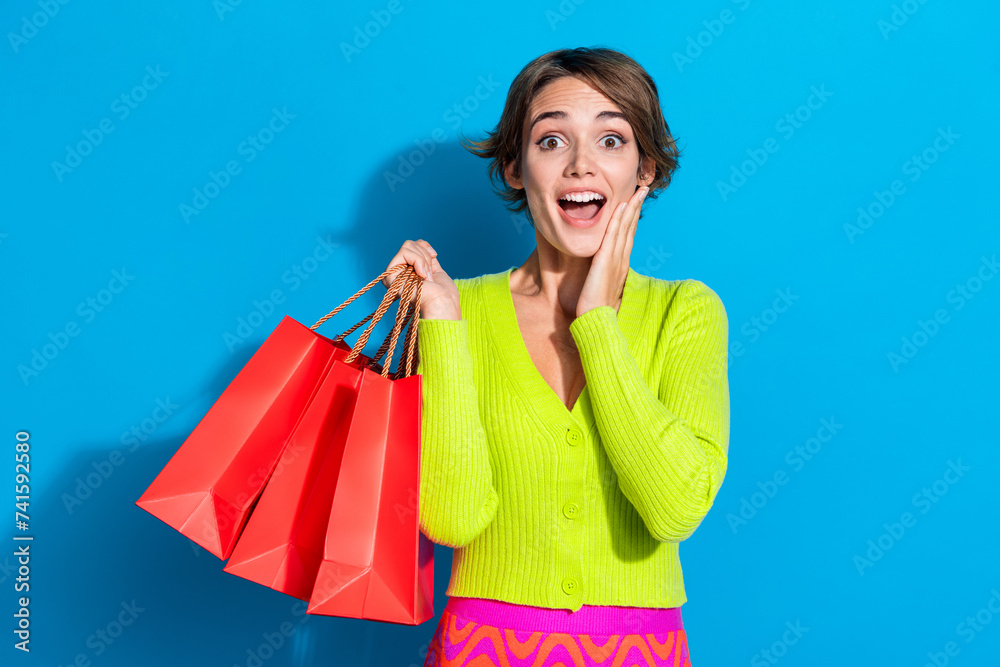 Sticker Portrait of young shopaholic lady in green cardigan touch cheek amazed holding gift boxes for birthday isolated on cyan color background