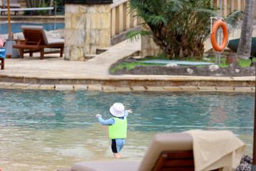 back view of unrecognized little boy playing in swimming pool wearing life vest and sun hat, concept of child safety while on holiday outdoors.