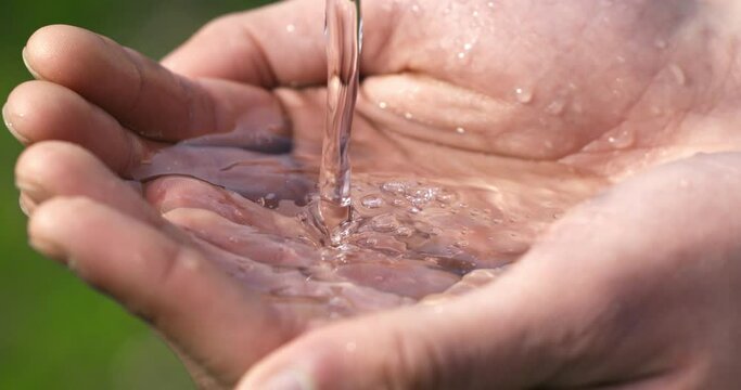 Super slow motion macro of crystal clear pure water flow stream pouring to young man hand with splashing drops on green nature background. Water saving, clean renewable energy and planet preservation.