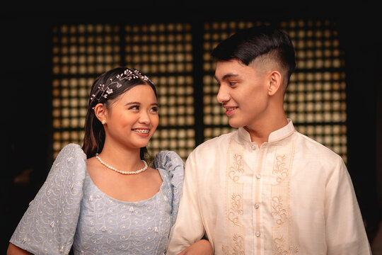 A young adorable couple showcases the beauty of Filipino traditional clothing, with a woman in a Terno and a man in a Barong Tagalog.