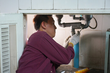 A male plumber repairs a pipeline or drain under the sink in kitchen room.