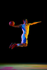 Dynamic photo of doing athlete man, basketball player doing perfect slam dunk in action against black studio background in neon light. Concept of professional sport, energy, match, tournament. Ad