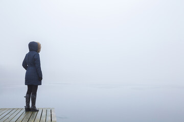Young adult woman standing on edge of footbridge and staring at lake in cold day. Mist over water....