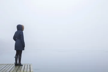 Fototapeten Young adult woman standing on edge of footbridge and staring at lake in cold day. Mist over water. Foggy air. Peaceful atmosphere. Spending time alone in nature. Empty place for text. Back view. © fotoduets