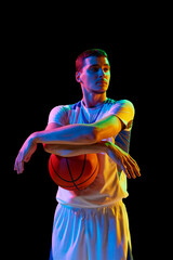 Portrait of confident young basketball player posing with ball against black studio background in mixed neon light. Concept of professional sport, energy, strength and power, match, tournament.
