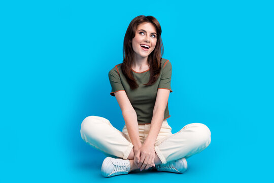 Full size photo of impressed woman wear green t-shirt sit on floor dreamy look at promo empty space isolated on blue color background