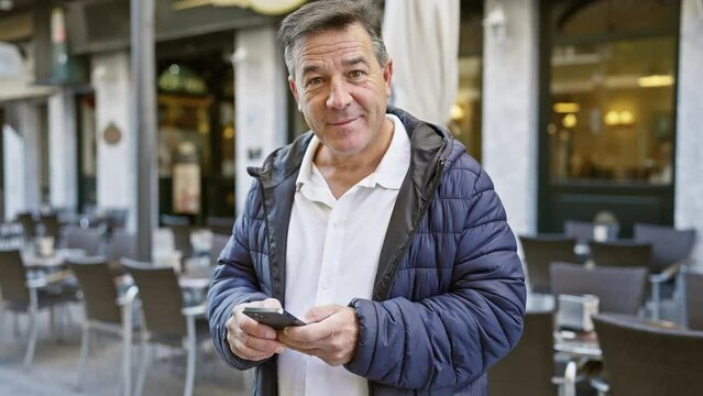 A smiling middle-aged man in a jacket using a smartphone at a city cafe terrace.