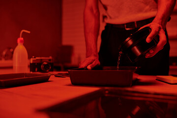 cropped view of photographer pours developer into tray for film processing in red darkroom