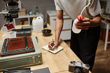 cropped dark skinned photographer prepares chemicals for film development and taking notes