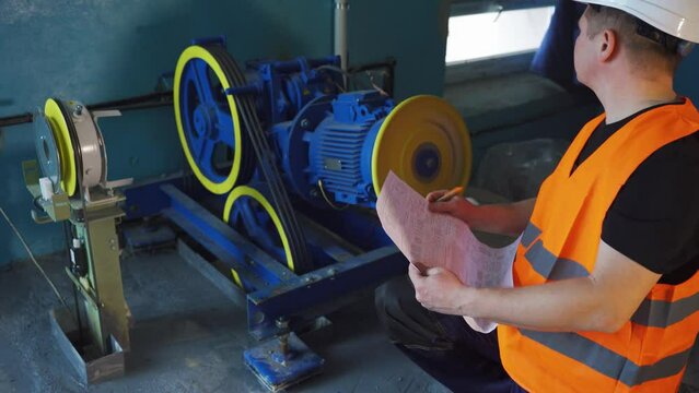 Launch and technical inspection of elevator equipment after repair. Engineer, worker in a white helmet, checks the performance indicators of an electric elevator motor in an old residential building