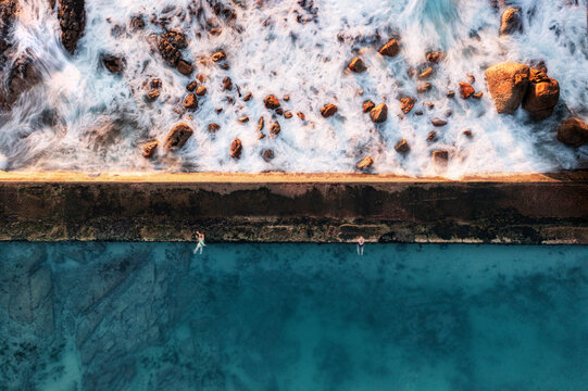 Aerial view of a tidal rock pool, taken with a long exposure shutter setting, showing the waves crashing onto the rocks and two people swimming in the pool, Cape Town, Western Cape, South Africa.