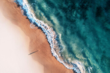 Aerial view of a white sandy beach with white waves rolling onto shore and crystal clear blue water and a girl standing on the beach casting a long shadow, Port Noarlunga, South Australia, Australia.