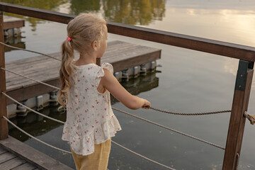 A kid, girl stands at parapet on wooden embankment of the lake or river looking at the water.  Peaceful natural landscape in summer, sustainable tourism. Safety fencing - Powered by Adobe
