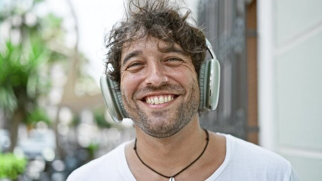 A cheerful man wearing headphones stands smiling on a sunny urban street, embodying casual style and happiness.