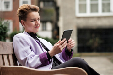 Young alternative woman reading an e-reader sitting on a bench under the rain.