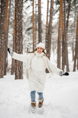 A cute young girl in a white jacket and hat runs and has fun in the winter in the forest, the girl has a good time outdoors, active lifestyle