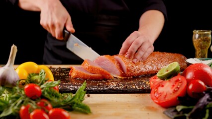 Chef cuts fresh meat on a wooden board on a dark background with vegetables and spices for marinade. Recipes and fresh produce concept.