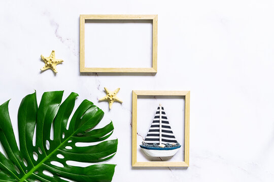 Summer vacation concept. Artistic composition with big tropical monstera leaf, decorative star fish and  wooden picture frame with a boat on white marble background. Copy space for your design.