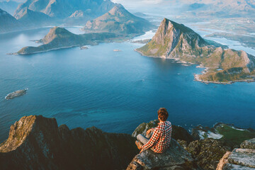 Man traveler sitting on the edge of mountain cliff in Norway alone hiking in mountains outdoor active healthy lifestyle adventure vacations tourist exploring Lofoten islands, aerial sea view - 741702561