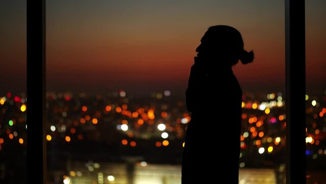 Man engages in business call at night, silhouette visible against window with city lights in background. Scene captures intense and busy urban lifestyle, showcasing professional in late-hour