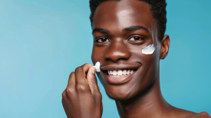 young man with a smile on his face applying a white cream on his cheek with his hand. - obrazy, fototapety, plakaty