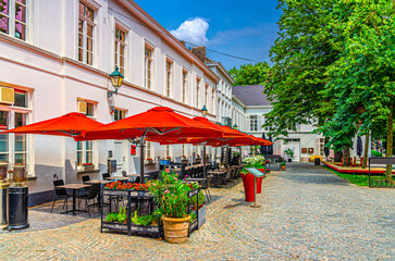 Street restaurant with tables, chairs and umbrellas near old building in stone paved courtyard in Kortrijk city historical centre in sunny summer day, Kortrijk old town, Flemish Region, Belgium