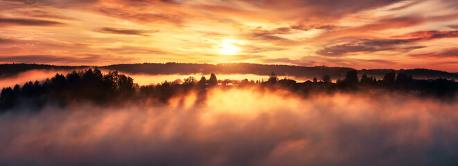 Dramatic sunrise over the fog and a range of tree silhouettes on a hill, a landscape panorama with dreamy sky, the rising sun and warm colors