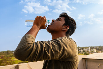 young man drinking water from plastic bottle on roof of house with blue sky