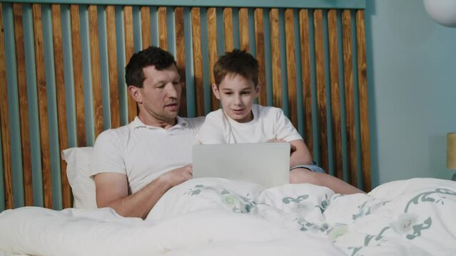 A friendly father and son in a joyful mood lie on the bed in the bedroom with a laptop, smile, laugh and talk. A positive dad with child watch a funny movie or cartoon and discuss it. The concept of
