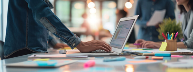 close-up view of a person's hands working on a laptop with other individuals in the background, surrounded by colorful sticky notes, papers, and a collaborative work environment.