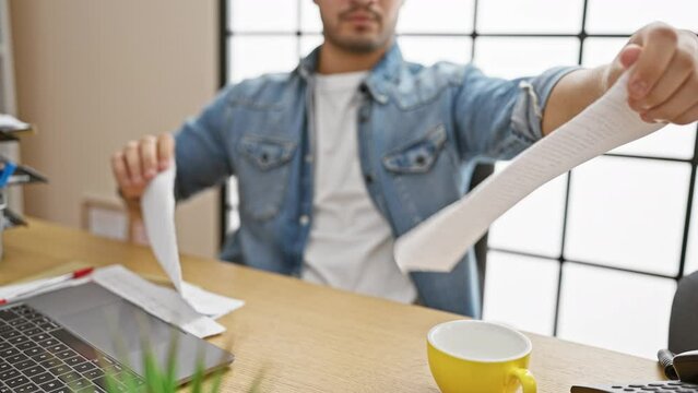 A man in an office tearing a document at a wooden desk with a laptop and coffee cup.