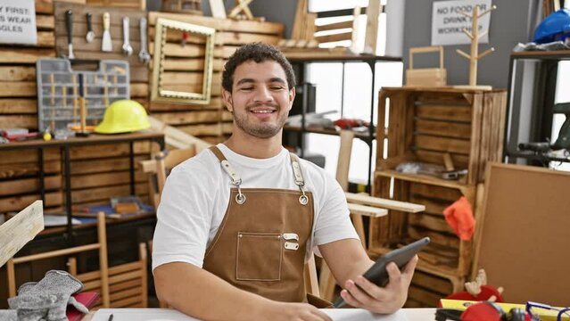 A smiling young man with a beard in a carpentry workshop wearing an apron using a smartphone, conveying a relaxed atmosphere.