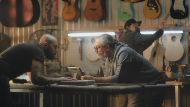 Mature male craftsman looks at laptop and uses digital tablet computer to draw blueprints of guitar. Tattooed carpenter holds electro guitar body and measures it using caliper in stylish workshop.