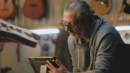 Close up of mature male carpenter using digital tablet computer to draw blueprint of guitar. Tattooed artisan talks with colleague and measures guitar body using caliper in light stylish workshop.