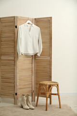 Stylish white shirt hanging on wooden folding screen, stool and boots indoors
