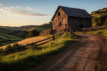 The warm evening sun bathes an old wooden granary and fence in golden light, with rolling hills stretching into the distance - obrazy, fototapety, plakaty