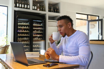Latin young man working on his laptop from a coffeeshop, on a lunch break, having croissant and coffee. Freelancing hispanic guy having a snack. Close up, copy space, background.