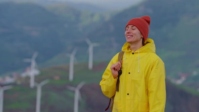 Attractive traveling woman in a yellow jacket and backpack looking at windmills in field 