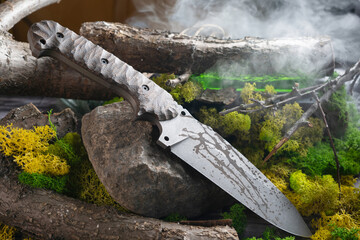 Military tactical knife lie on nature background. Stone, moss, trunk and wooden twigs in the forest. Mystique background with fog