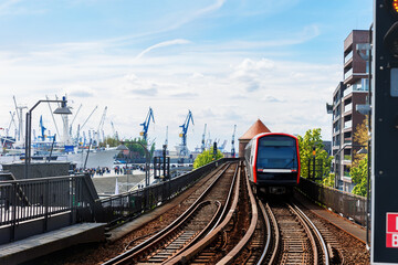 Hamburg Red U-bahn Subway train departure Baumwall station on Elevated Track with Hamburger Port Cranes and ships cargo harbor background blue sky. Hanseatic city commute transport scenic view