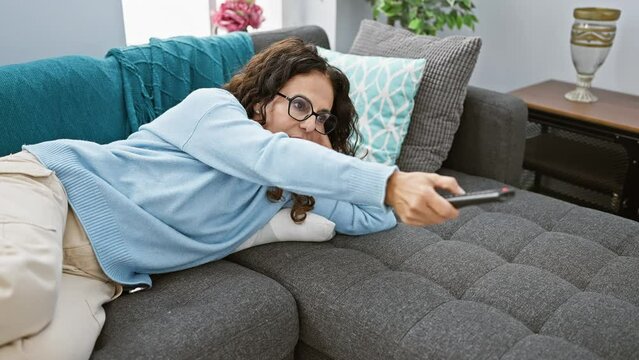 Hispanic woman lounging on sofa in living room with remote control, glasses, smiling, relaxed, casual, comfortable, leisure, lifestyle, interior