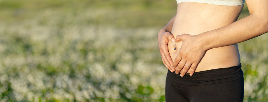 Pregnant woman in the field of flowers in the first trimester forming a heart with her hands on her belly with a piercing in her navel and blurred background for copy. Pregnant woman with flowers