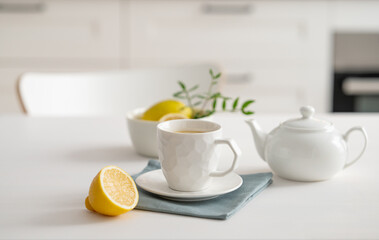 A cup of tea with lemon and a teapot on a white table against the background of a white kitchen.