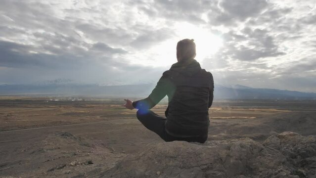 Rear view of a young man with a ponytail sitting in the lotus position on a stone meditating at sunset in the mountains. Practicing yoga alone and calm. Spiritual balance and harmony with nature